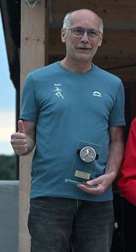 Ein älterer Mann in einem blauen T-Shirt hält eine Trophäe in der Hand, gibt ein Daumen hoch, mit einer Holzstruktur und klarem Himmel im Hintergrund.