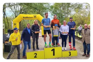 Eine Gruppe von Menschen steht auf einem Podium mit Trophäen, ein Mann hält ein Mikrofon und ein gelber Banner mit dem Wort Raiffeisen steht dahinter.