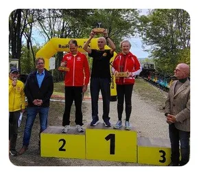 Eine Gruppe von Menschen steht auf einem Podium mit den Nummern 1, 2 und 3. Sie halten Trophäen und scheinen einen Sieg zu feiern. Im Hintergrund ist ein gelber Bogen und Bäume zu sehen.