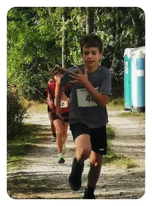 Ein junger Junge in einem grauen T-Shirt mit der Nummer 4 darauf läuft in einem Rennen. Hinter ihm nehmen auch zwei andere Läufer an dem Rennen teil.