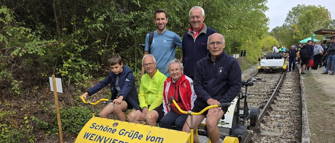Eine Gruppe von Menschen fährt mit einem kleinen gelben Zug auf einer Schiene. Der Zug hat ein Schild mit der Aufschrift 'Schone Gruße vom Weinviertel-Express!'.