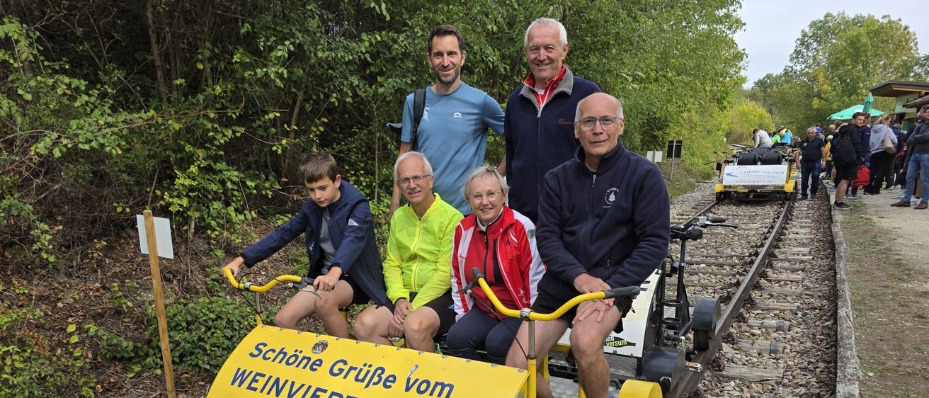 Eine Gruppe von Menschen fährt mit einem kleinen gelben Zug auf einer Schiene. Der Zug hat ein Schild mit der Aufschrift 'Schone Gruße vom Weinviertel-Express!'.