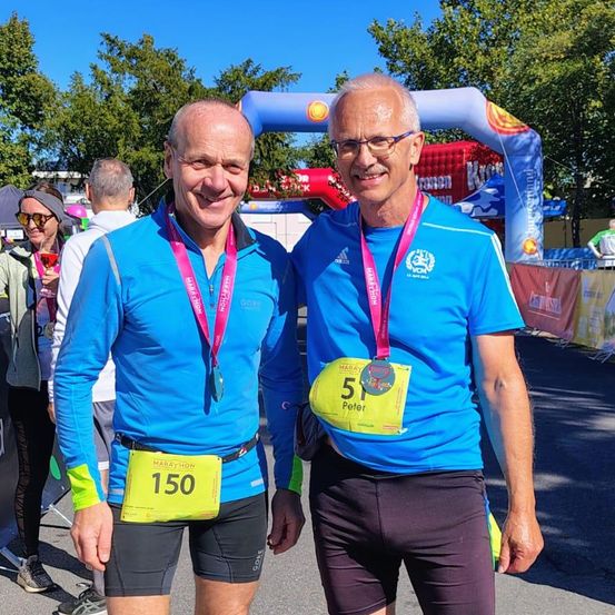 Zwei Männer mit Marathon-Medaillen posieren für ein Foto bei einer Marathon-Veranstaltung. Beide tragen blaue Shirts, schwarze Shorts und Sneakers. Der Mann auf der linken Seite hat eine Medaille mit der Nummer 150. Dahinter sind Zelte, Bäume und eine Menschenmenge.
