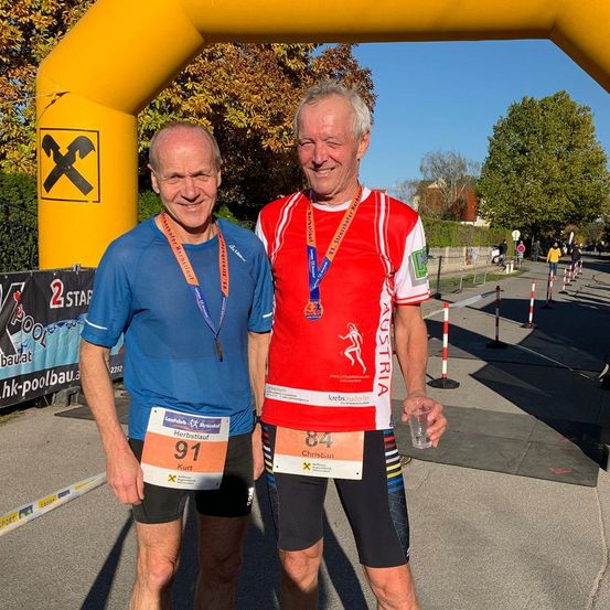 Zwei Männer stehen lächelnd vor einem gelben Bogen mit Raiffeisen darauf. Sie tragen beide Marathonnummern. Einer trägt Blau und der andere Rot. Hinter ihnen befindet sich eine Straße mit mehreren Menschen, die gehen.