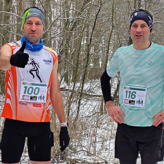 Zwei Männer in Laufausrüstung stehen in einem verschneiten Wald. Ein Mann gibt einen Daumen hoch und trägt die Nummer 100 auf seinem Shirt. Der andere Mann hält seine Brust und trägt die Nummer 116.