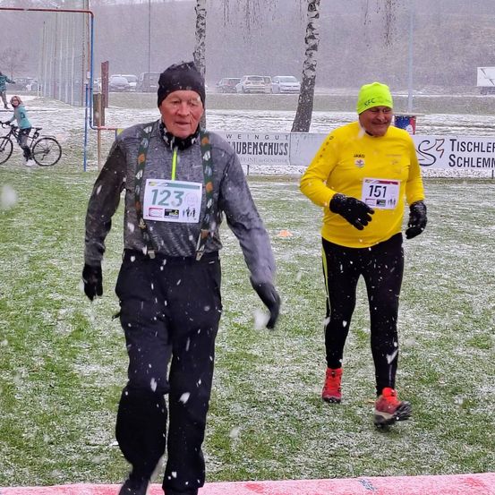 Zwei Männer in warmer Kleidung laufen auf einem verschneiten Feld. Einer trägt ein gelbes Shirt mit der Nummer 151, der andere ein graues Shirt mit der Nummer 123. Hinter ihnen steht ein Zaun und ein Baum mit einem Schild.