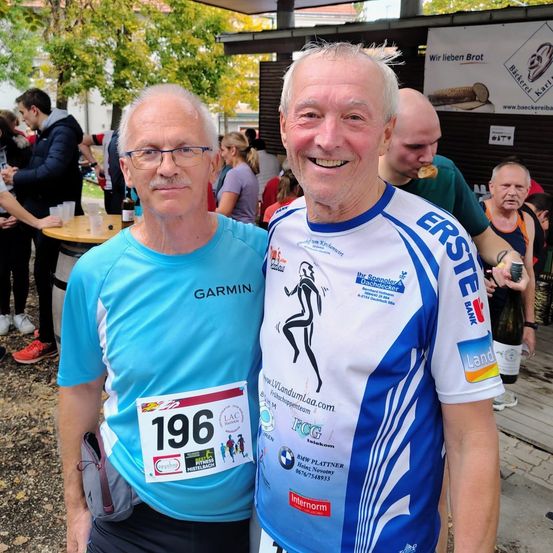 Zwei ältere Männer in Laufbekleidung stehen lächelnd nebeneinander in einem überfüllten Bereich. Hinter ihnen stehen Menschen, die an Tischen essen und trinken. Ein Mann trägt ein Shirt mit der Nummer 196.