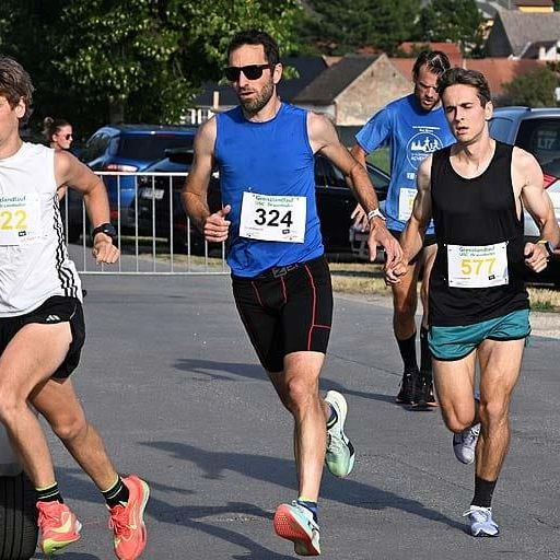 Drei Läufer sprinten bei einem Marathonrennen. Der Mann vorne trägt die Nummer 522, gefolgt von einem Mann in Blau mit der Nummer 324 und einem anderen in Schwarz mit der Nummer 577. Sie sind von geparkten Autos und Bäumen umgeben.