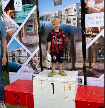 Ein junger Junge steht auf einem Podium mit der Nummer eins darauf, hält eine Trophäe und einen Beutel. Er trägt ein Fußballtrikot und Turnschuhe. Hinter ihm befinden sich zwei Banner mit Bildern von Gebäuden und einer Person.