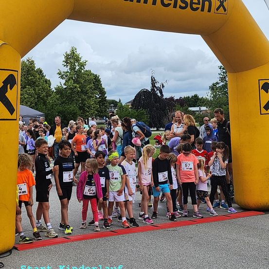 Eine Gruppe von Kindern und Erwachsenen steht an der Startlinie eines Rennens, trägt nummerierte Startnummern und Turnschuhe, mit einem gelben Bogen im Hintergrund.