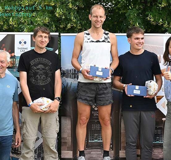Fünf Personen stehen auf einer Bühne, lächeln und halten blaue Boxen in den Händen, mit einem Hintergrund, der ein Banner und Bäume zeigt. Der Mann in der Mitte trägt ein weißes Tanktop und Shorts.