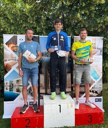 Drei Männer stehen auf einem Podest mit den Nummern 1, 2 und 3. Der Mann in der Mitte hält eine Medaille und ein Zertifikat, während die anderen Trophäen halten. Sie sind lässig gekleidet und stehen vor einer Kulisse mit Hotel-Werbung.
