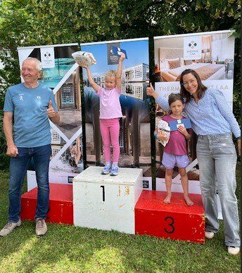 Drei Personen stehen auf einem Podest mit der Nummer 1 auf der mittleren Stufe. Ein Mann links zeigt Daumen hoch, ein Mädchen in Pink steht auf der mittleren Stufe und hält einen Pokal, und eine Frau rechts hält einen Pokal. Sie alle lächeln.