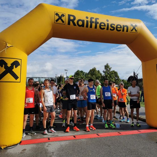 Eine Gruppe von Läufern mit nummerierten Startnummern steht unter einem gelben Bogen mit dem Wort Raiffeisen darauf. Sie nehmen wahrscheinlich an einem Marathon teil.