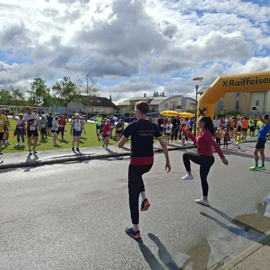 Eine Gruppe von Läufern nimmt an einem Marathon teil. Im Vordergrund sind ein Mann und eine Frau beim Dehnen zu sehen. Dahinter ist ein gelber Bogen mit dem Wort Raiffeisen sichtbar.