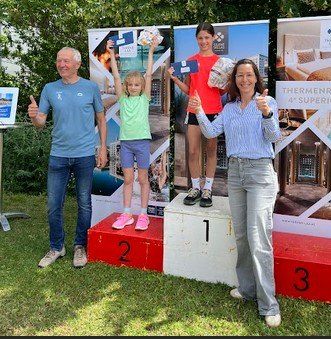 Drei Personen stehen auf einem Podest mit den Nummern 1, 2 und 3. Ein Mann in Blau, ein Mädchen in Grün und eine Frau in Weiß. Sie alle lächeln und geben Daumen hoch. Hinter ihnen sind Banner mit Bildern von Menschen und Gebäuden.