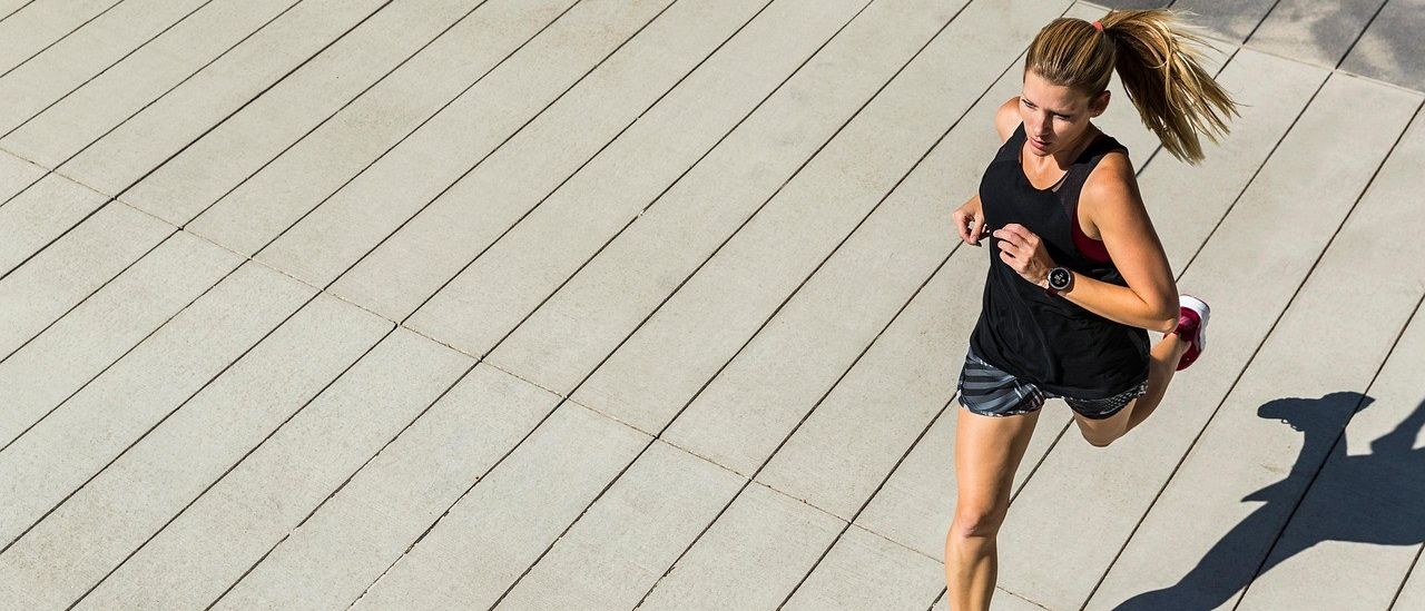 Eine Frau in einem schwarzen Tanktop und roten Turnschuhen läuft auf einem Holzweg mit einem Schatten auf dem Boden.