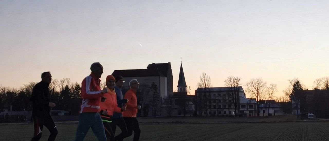Vier Personen joggen auf einem Weg in der Nähe eines Feldes. Dahinter steht ein großes Gebäude mit einem Kirchturm und einem klaren Himmel.