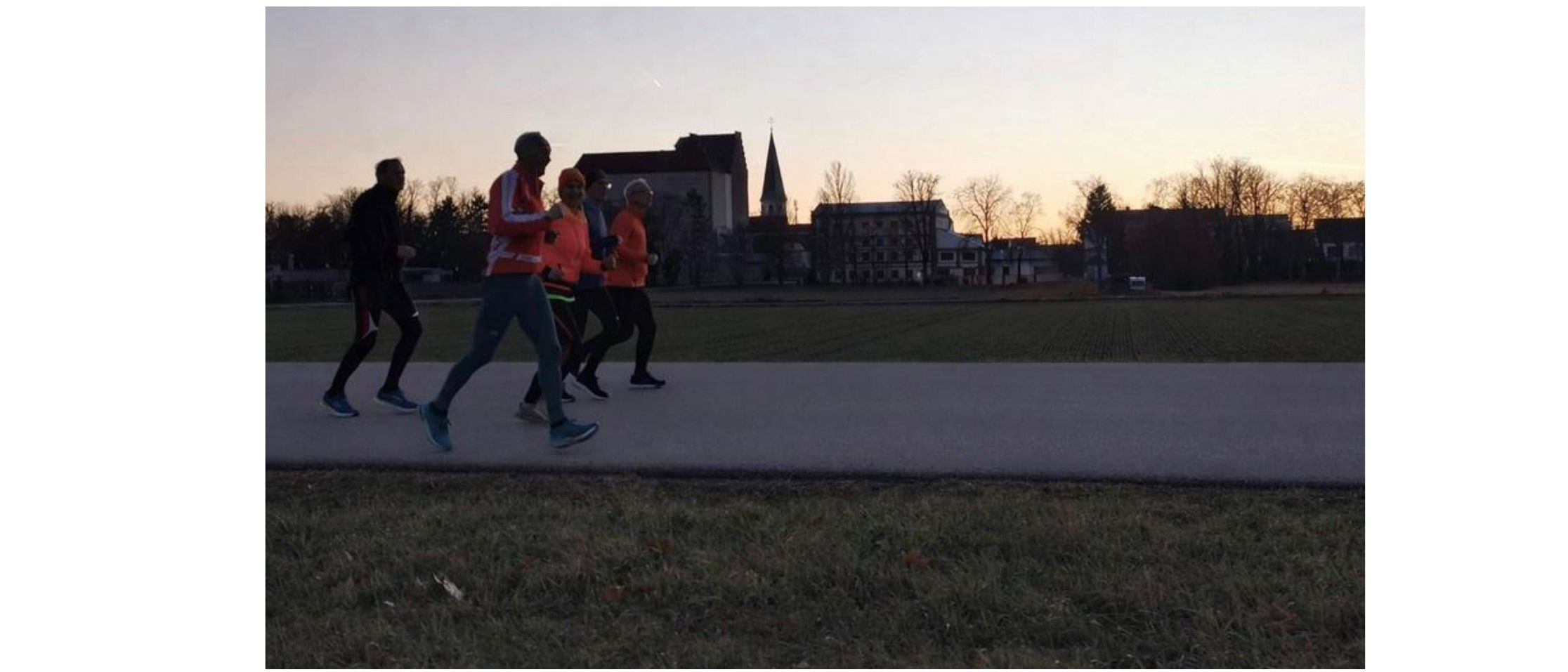 Eine Gruppe von Menschen joggt auf einem von Bäumen und Gebäuden umgebenen Pfad während eines Sonnenuntergangs.