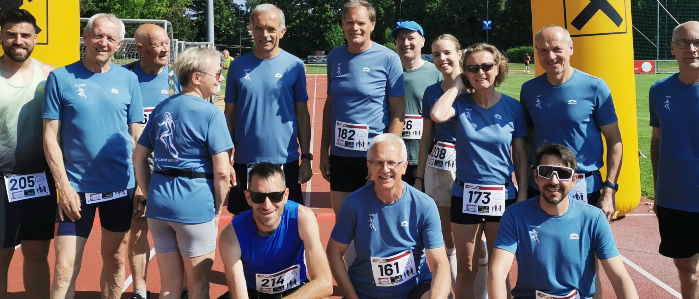 Eine Gruppe von Menschen in blauen Shirts und mit Sonnenbrillen posiert für ein Foto auf einer Laufbahn. Sie sind wahrscheinlich Teilnehmer eines von Raiffeisen gesponserten Marathons.