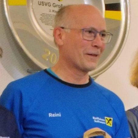 Ein Mann in einem blauen Shirt mit dem Wort Reini hält einen braunen Papierbeutel. Auf dem Beutel ist ein Logo und er lächelt.