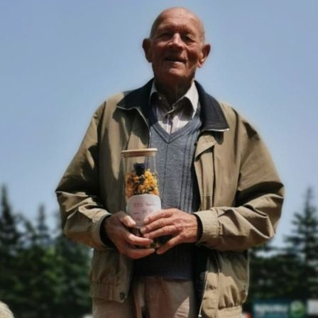 Ein Mann steht auf einem Rasenfeld und hält eine Dose mit Essen. Eine Frau sitzt neben ihm und beobachtet. Bäume und ein Gebäude sind im Hintergrund.