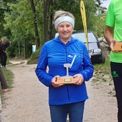 Eine Frau steht auf einem Podest und hält eine Trophäe in der Hand, neben ihr steht ein Mann, beide lächeln. Hinter ihnen ist ein Auto in der Nähe eines gelben Banners geparkt, und Menschen stehen auf einem Weg, der von Bäumen umgeben ist.