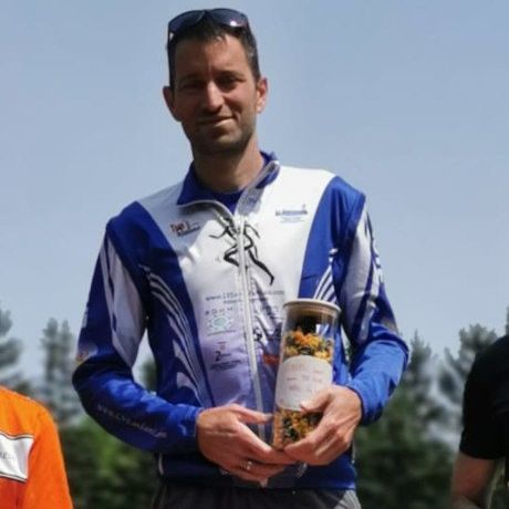 Ein Mann steht auf einem Podest und hält ein Glas mit gelben Blumen. Dahinter stehen zwei andere, einer in Orange und der andere in Schwarz. Sie befinden sich auf einem Feld mit Bäumen und einem Gebäude in der Ferne.