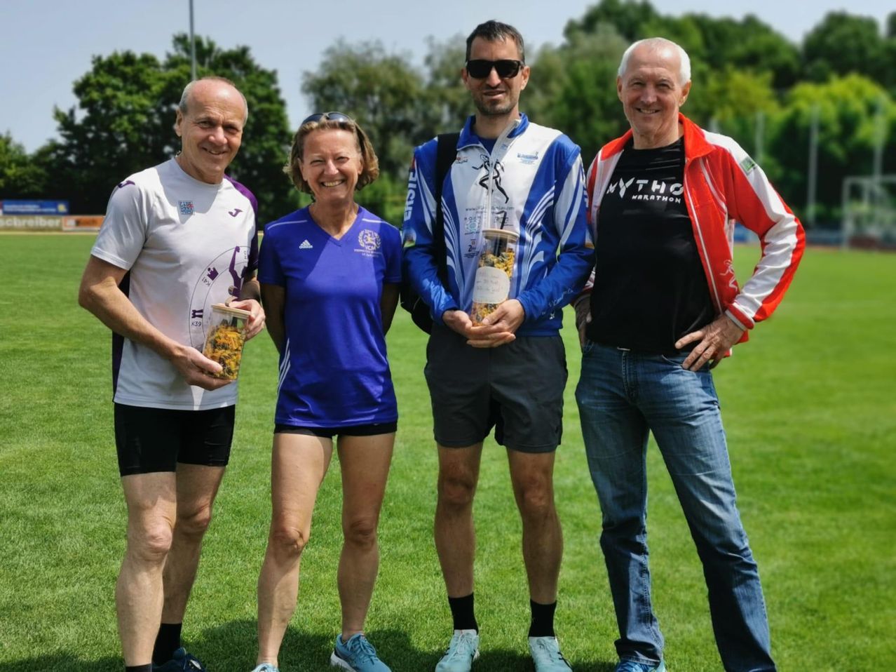 Vier Personen stehen auf einem Feld und lächeln. Der Mann auf der linken Seite hält einen Pokal, und die Frau in der Mitte lächelt breit. Der Mann neben ihr hält einen Pokal mit einem gelben Objekt darin. Der Mann auf der rechten Seite trägt eine Jacke mit 'Mytho Marathon' darauf. Sie alle tragen Turnschuhe. Bäume und ein Pfosten sind im Hintergrund.