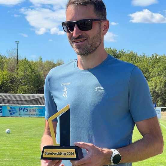 Ein Mann in einem blauen Shirt mit Logo hält einen Pokal auf einem Fußballfeld.