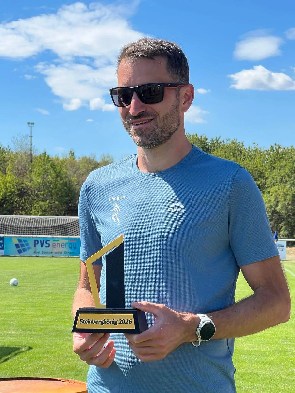 Ein Mann in einem blauen Shirt mit Logo hält einen Pokal auf einem Fußballfeld.