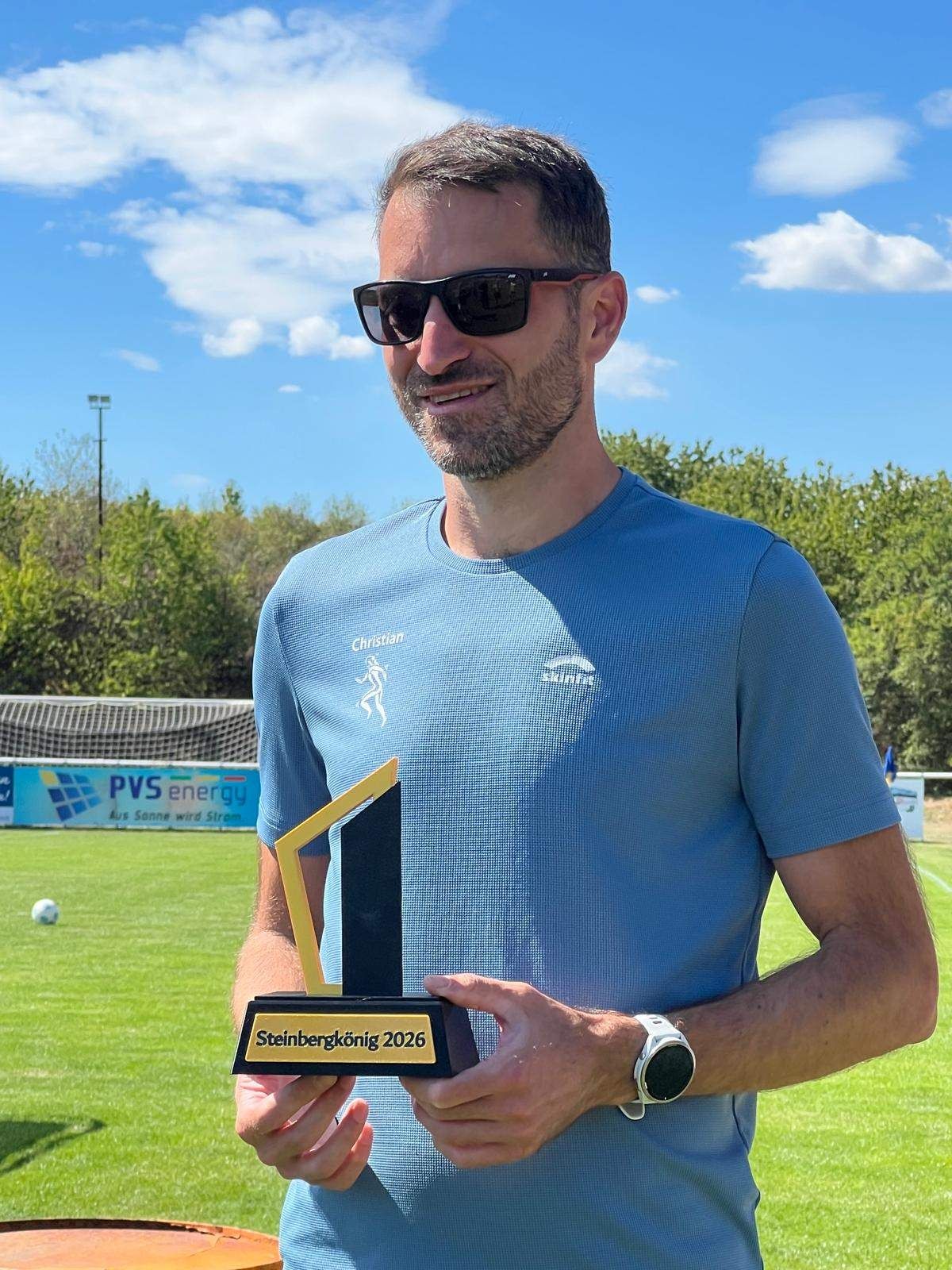 Ein Mann in einem blauen Shirt mit Logo hält einen Pokal auf einem Fußballfeld.