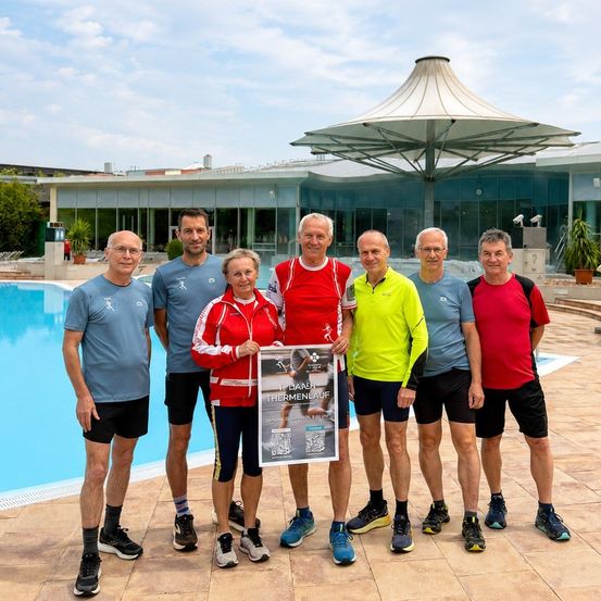 Eine Gruppe älterer Männer und Frauen steht vor einem Pool und hält ein Poster für den 10. Diacher Thermenlauf.