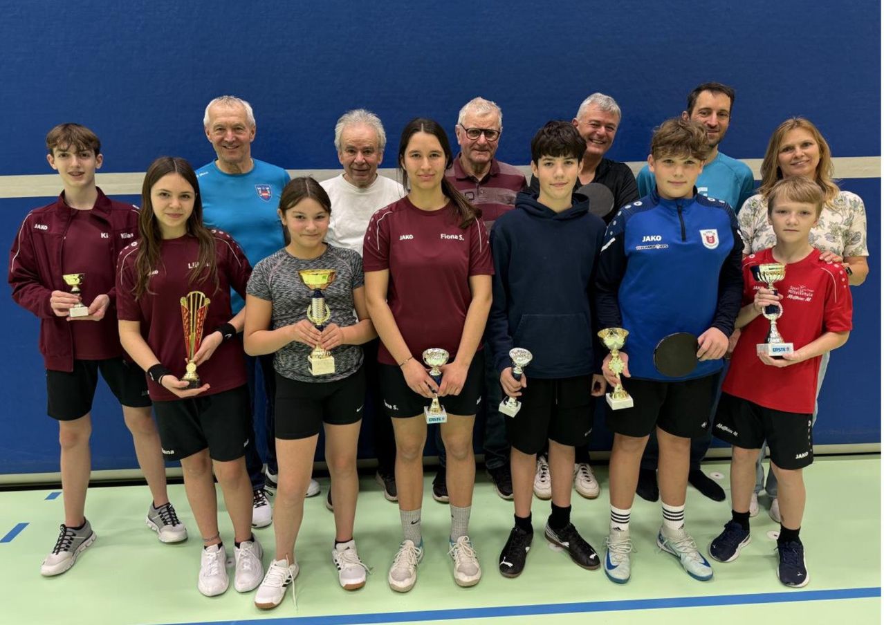 Eine Gruppe von Menschen, wahrscheinlich ein Sportteam, steht in einer Reihe mit einer blauen Wand im Hintergrund. Sie halten Pokale und einige haben Schläger.