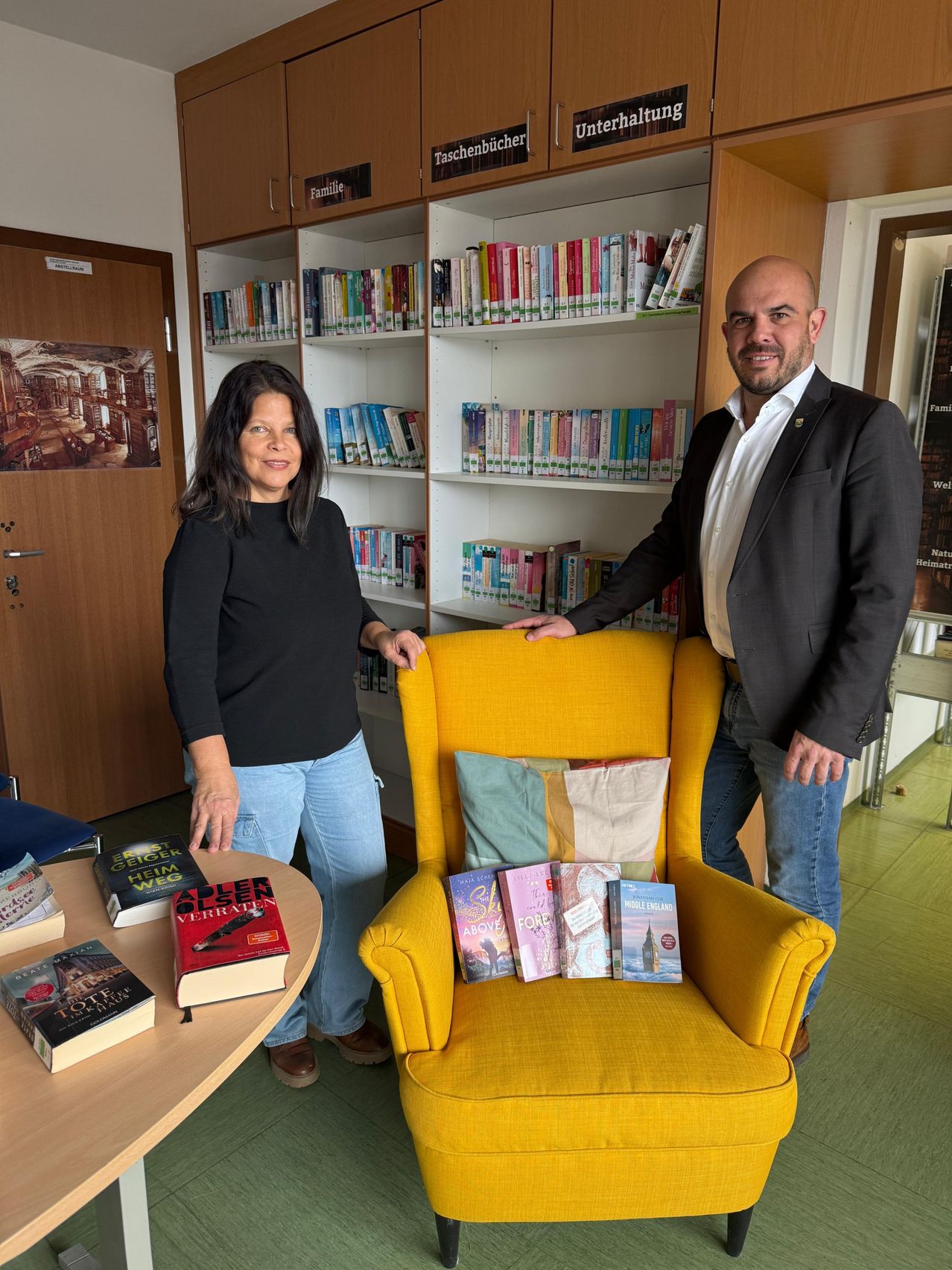 Zwei Personen stehen in einer Bibliothek. Eine Frau steht neben einem Tisch mit Büchern darauf. Ein Mann steht hinter einem gelben Stuhl mit einem Stapel Bücher darauf. Sie lächeln beide. Hinter ihnen stehen Regale voller Bücher.