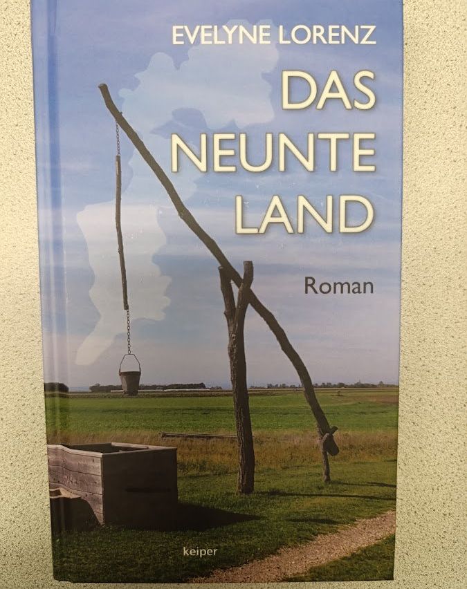 Buchcover mit dem Titel 'Das Neunte Land' von Eveline Lorenz. Es zeigt ein Feld mit einem hölzernen Brunnen und einem Eimer, der an einem hölzernen Hebel hängt.