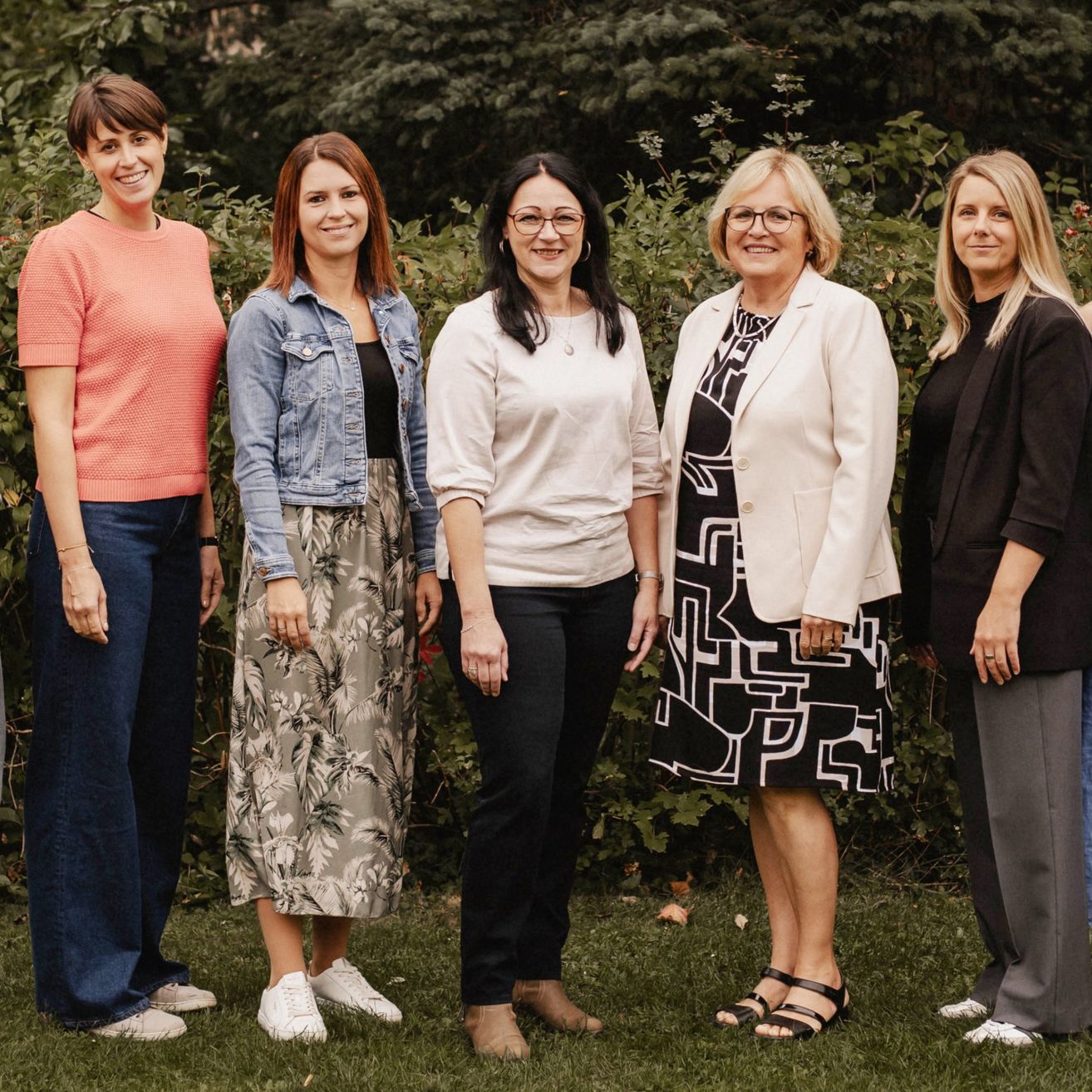 Fünf Frauen stehen lächelnd in einem Garten, gekleidet in lässige bis formelle Kleidung. Im Hintergrund ist üppige Vegetation zu sehen.