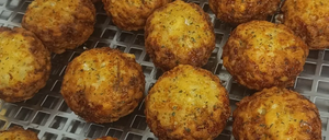 Nahaufnahme von golden-braunen, knusprigen, runden Kartoffelbällchen mit Käse und Kräutern auf einem Metallgitter.