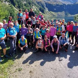 Eine Gruppe von Wanderern posiert für ein Foto vor einem Bergsee. Sie tragen Rucksäcke, Sonnenbrillen und Wanderausrüstung. Der See ist von Bäumen und Bergen umgeben.