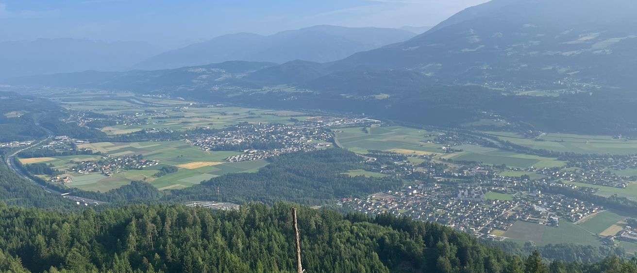 Luftaufnahme eines Tals mit einer Stadt unten, umgeben von grünen Feldern und Bergen im Hintergrund. Der Himmel ist klar mit einigen Wolken.