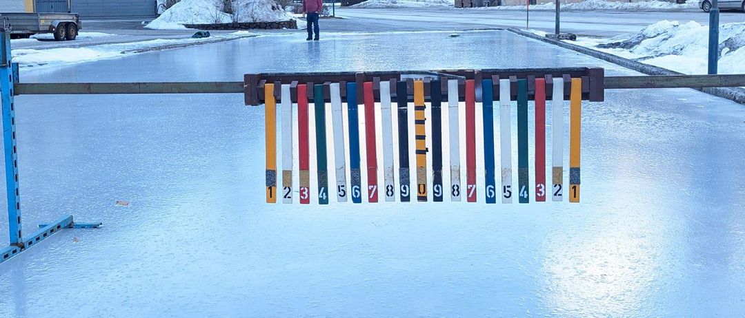 Eine vereiste Eislaufbahn mit nummerierten Stangen in einer verschneiten Bergstadt. Eine Person steht in der Ferne.