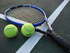 Ein Tennisschläger und zwei Tennisbälle liegen auf einem Tennisplatz. Der Schläger hat einen blauen Griff und schwarzes Band. Die Bälle sind gelb mit weißen Schriftzügen.
