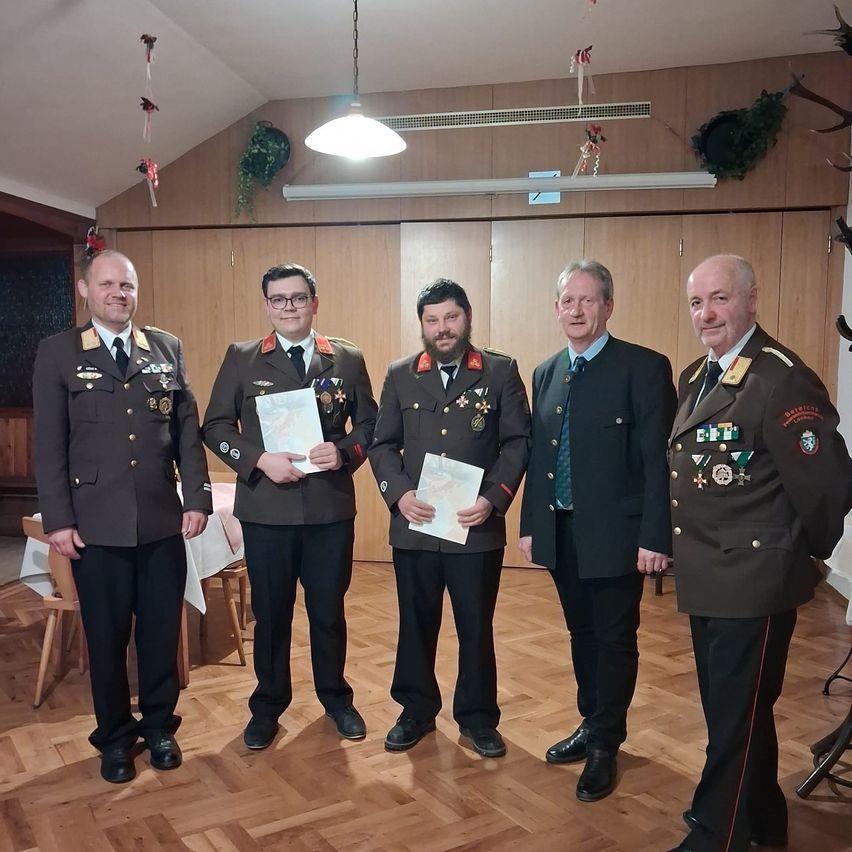 Fünf Männer in Militäruniformen stehen in einem Raum. Einer hält ein Zertifikat. Holzwände, eine Lampe und Dekorationen sind im Hintergrund.