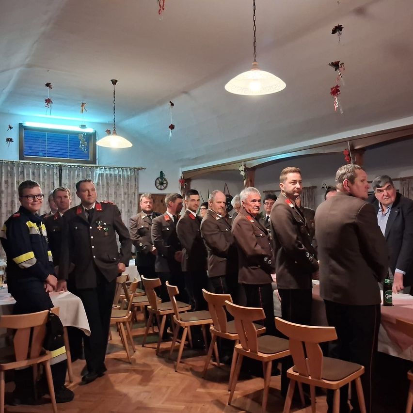 Mehrere uniformierte Männer stehen in einer Reihe, möglicherweise bei einer formellen Veranstaltung. Einige tragen Hüte und Brillen. Hinter ihnen steht ein Tisch mit Stühlen, und ein Mann hält eine Dose.