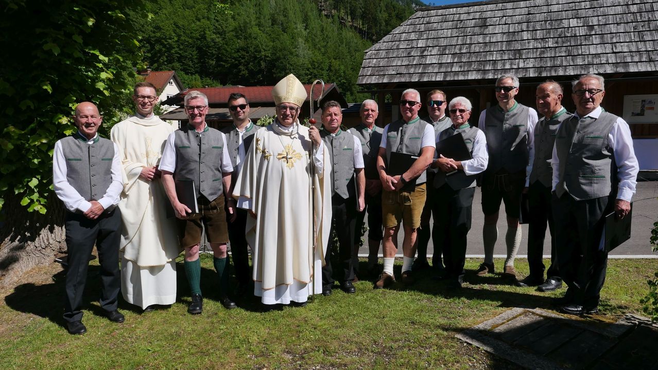 Ein Priester mit einem goldenen Kreuz auf der Brust steht mit einer Gruppe von Männern in traditioneller Kleidung und posiert möglicherweise für ein Foto.