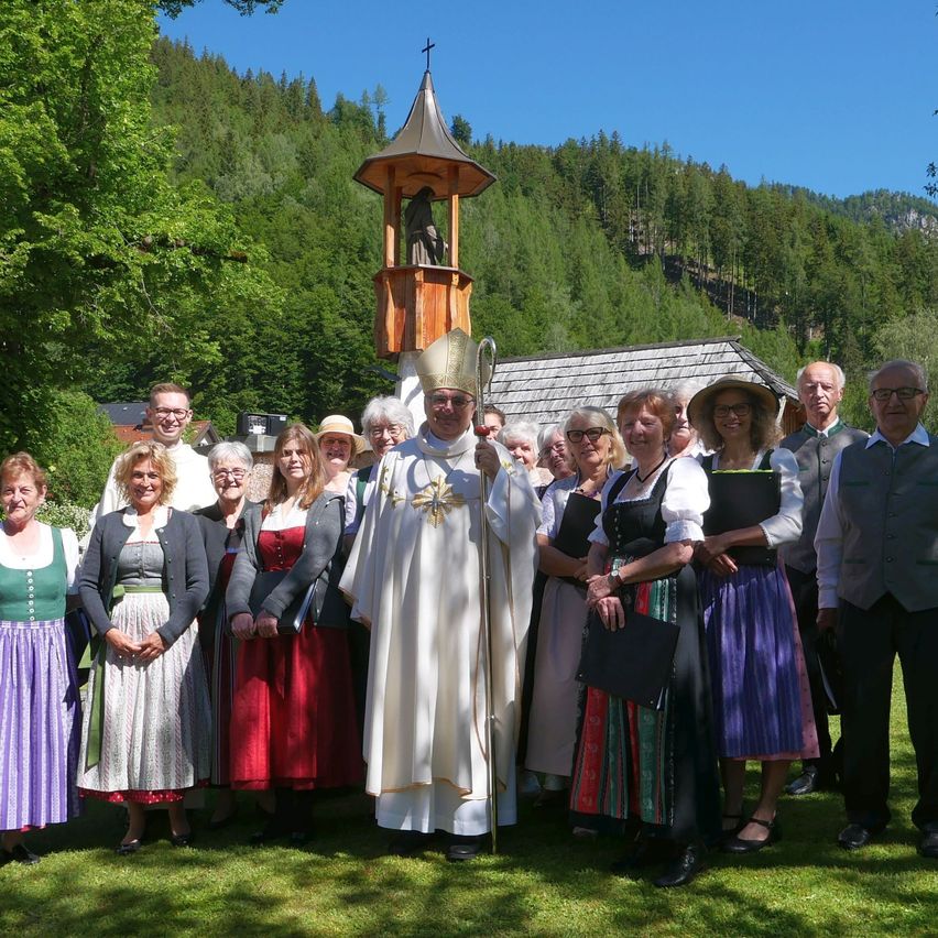 Eine Gruppe von Menschen in traditioneller Kleidung steht mit einem Priester vor einer kleinen Kapelle mit Glockenturm in einem bergigen Gebiet.