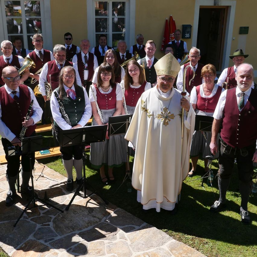 Ein Priester steht mit einem Chor und einer Band, einen Stab in der Hand, auf einem Rasen vor einem gelben Gebäude. Sie tragen traditionelle Westen und Gewänder.