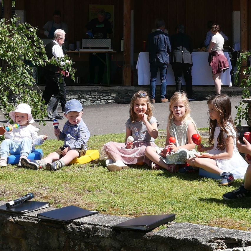 Kinder sitzen auf dem Gras und halten Maracas. Einige Erwachsene stehen hinter ihnen in der Nähe eines Tisches. Im Hintergrund ein Mann in einem weißen Hemd und einer Schürze.