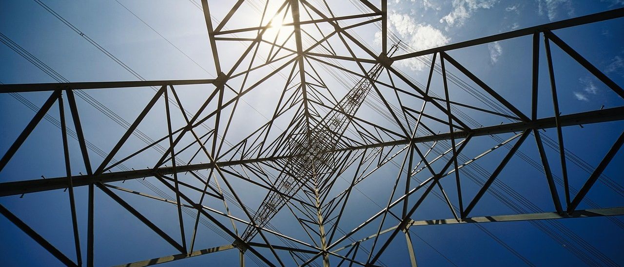 Ein Hochspannungsmast erhebt sich gegen einen blauen Himmel, in dem die Sonne hell scheint. Die Struktur besteht aus sich kreuzenden Metallträgern.