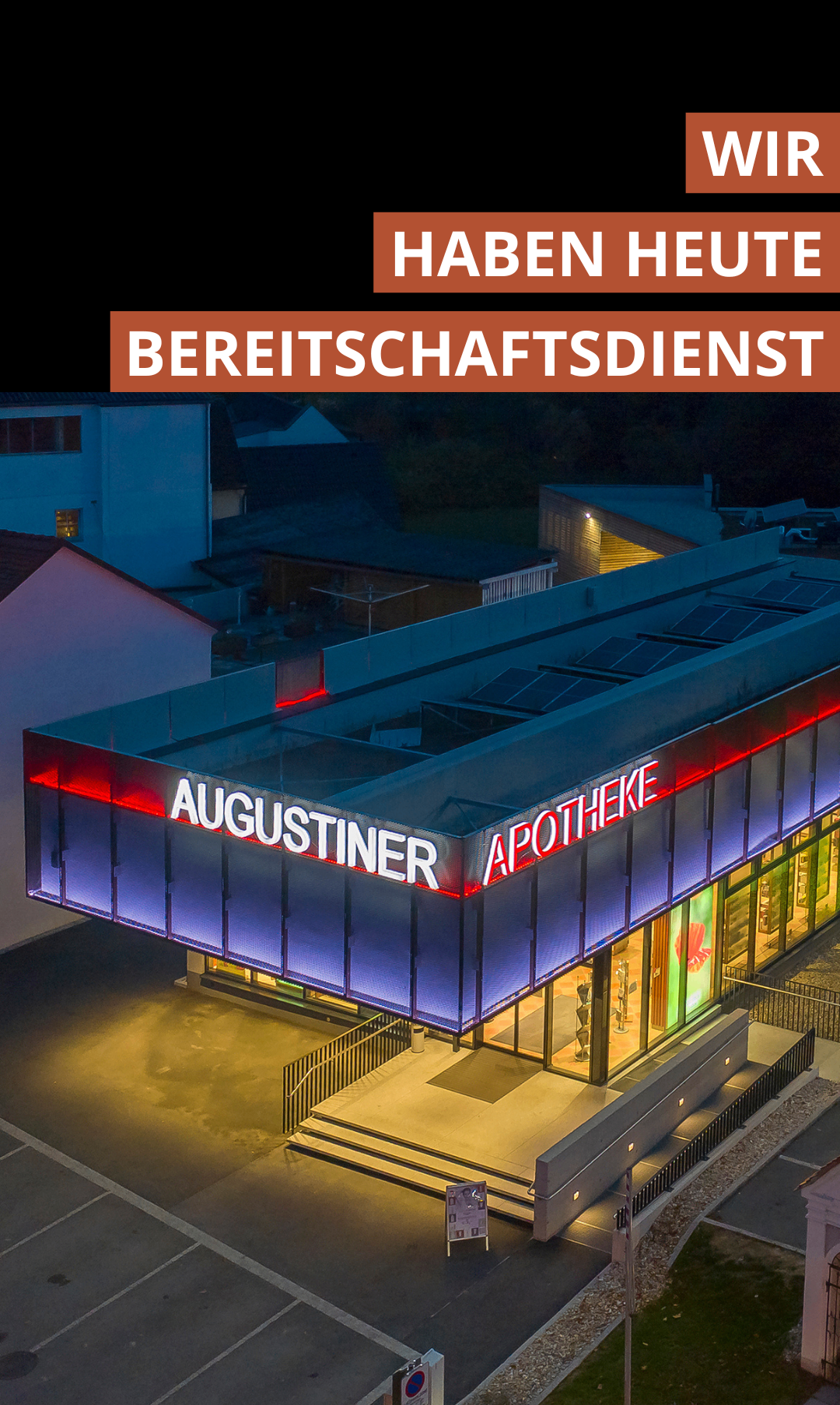 An evening view of a building, with the word 'AUGUSTINIER APOTHEKE' in large white letters on the side. The building has solar panels on its roof and is brightly lit up.